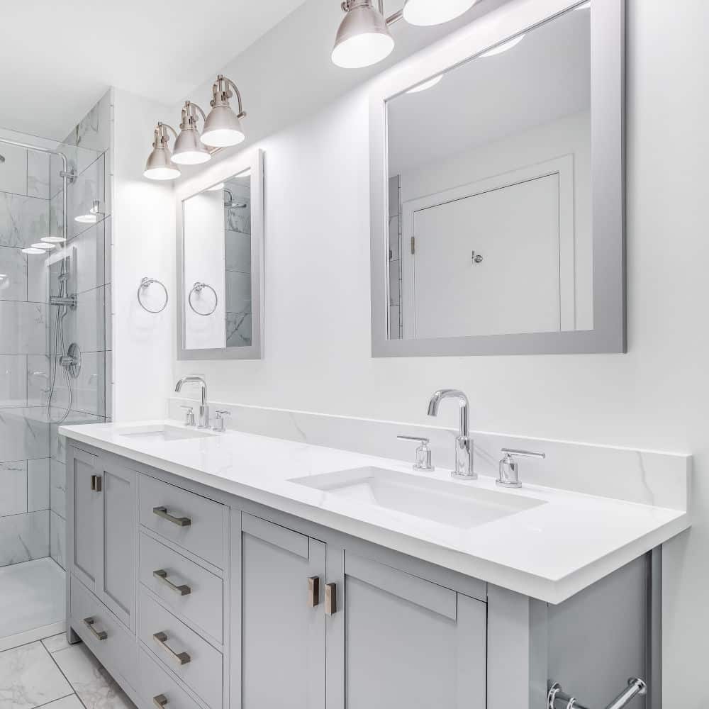 An elegant, remodeled bathroom with a grey vanity and bronze hardware. The shower has a large shower head and marble tiles and glass wall line the sides.